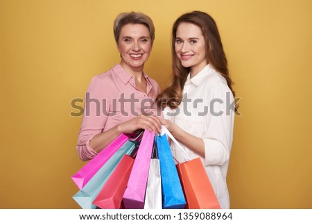 Cropped photo of stylish smiling ladies wearing fashionable blouses and making shopping. They are posing against yellow background with colorful paper bags in arms. Black friday sale concept