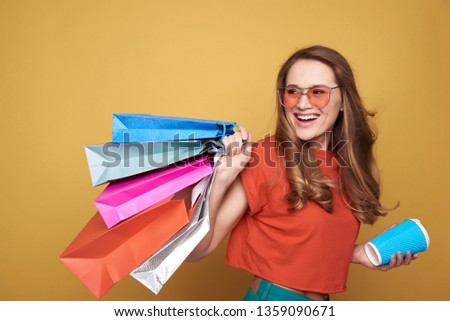 Waist up of happy Caucasian brown-haired girl with beautiful hairstyle wearing fashionable eyeglasses. She is keeping shopping bags on shoulder and drinking coffee while looking away against orange