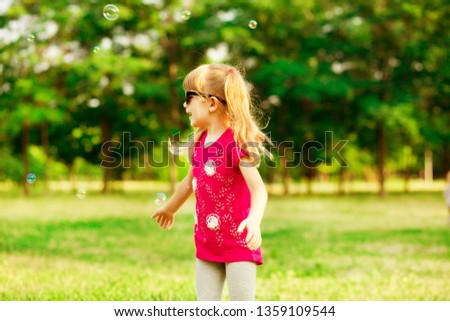 Happy little blond girl blows soap bubbles in city summer park