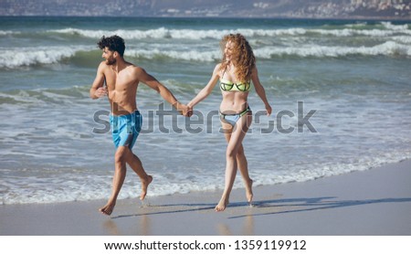 Front view of happy young Caucasian couple holding hands and running by the oean at beach on sunny day. They are smiling