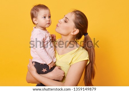 A young caring mother tries to calm down her crying daughter. A tear is going to run down cute baby`s cheek. A disappointed child looks concentrated on something aside. Mum kisses her amazing kid.