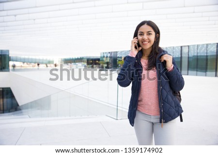 Cheerful Indian student girl talking on mobile phone. Portrait of happy young woman with satchel walking in city. Communication concept