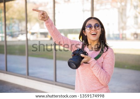 Joyful woman holding camera and pointing away outdoors. Pretty young lady looking at camera and standing with mirror glass wall in background. Photography concept. Front view.