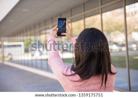 Back view of woman taking selfie photo outdoors. Young lady holding smartphone and standing with building wall and walkway in background. Selfie and tourism concept. Back view.