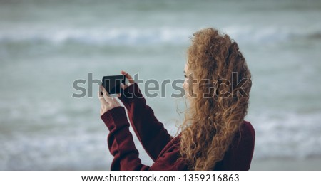 Side view of beautiful young Caucasian woman taking picture with her mobile phone standing at beach