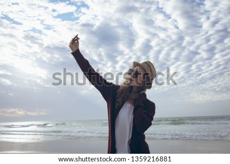 Side view of pretty young Caucasian woman with hat and sunglasses taking selfie with her mobile phone standing at beach.