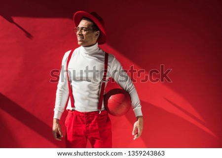 stylish mixed race man in hat and suspenders posing with basketball on red with copy space