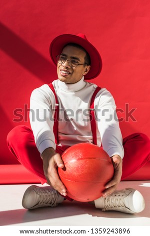 stylish mixed race man in hat and suspenders sitting, smiling and posing with basketball on red