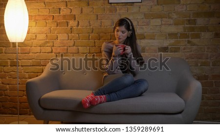 Closeup portrait of young attractive caucasian female listening to music in headphones while sitting on the couch and holding a cup of tea indoors
