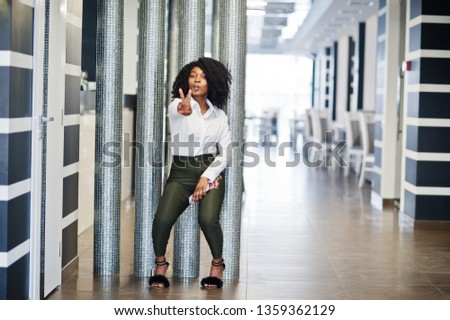 Сheerful business african american lady with afro hair, wear white blouse and green pants posed in cafe with mobile phone at hand show V finger sign.