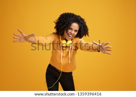 Lovely african american young woman with bright smile dressed in casual clothes and headphones ready for hugs over yellow background.