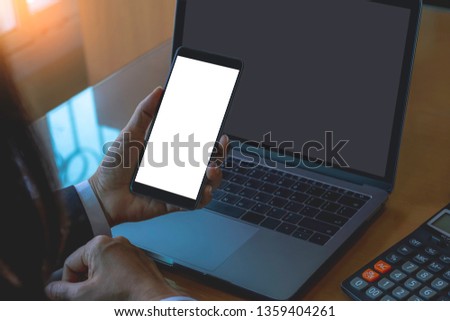 Mock up image of business woman hand holding and showing mobile smartphone with blank white screen, work on laptop computer and calculator on the table at office. Flare light.