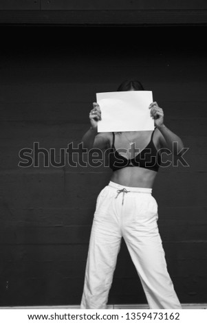 Caucasian female model holding up A4 paper, presenting A4 paper, covering her face. Girl shows blank paper. Black and white, high contrast image