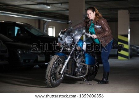 Woman biker trying to sit on motorcycle, standing in dark parking