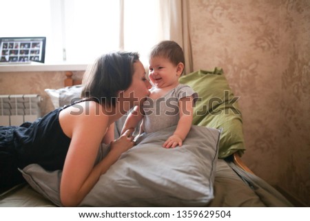 funny young mother brunette kisses and hugs the baby daughter, real bedroom interior, casual style