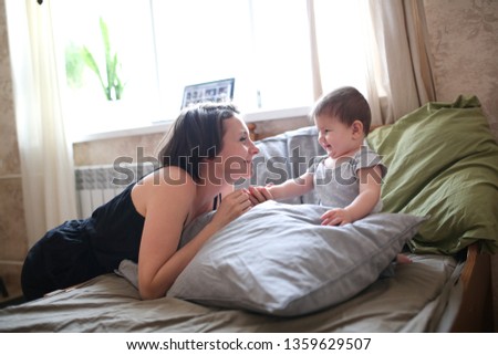 funny young mother brunette kisses and hugs the baby daughter, real bedroom interior, casual style