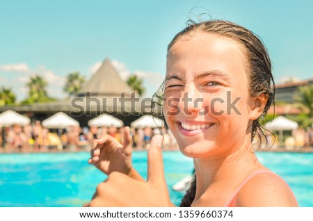 Smiling teen girl enjoing summer vacation at the hotel pool with palms and sun umbrellas on the background. Girl shows thumbs up symbol. Summer holidays, fun summertime and watersports concept.