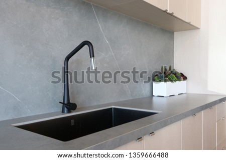 A black sink and a matt-finish black faucet set against a grey countertop and backsplash made of porcelain slabs that mimic the natural look of stones, decorated by green indoor planter. Royalty-Free Stock Photo #1359666488
