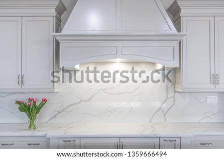 A modern white kitchen with a traditional touch. White marble-looking quartz countertop and backsplash accompanied by white cabinetry and custom made range hood. Royalty-Free Stock Photo #1359666494