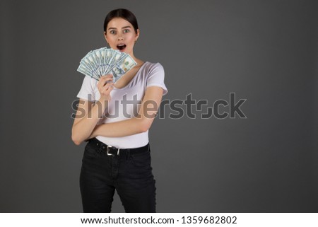 Portrait of young brunette woman in whote t-shirt holding money banknotes and celebrating isolated over grey background