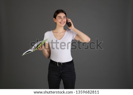 Angry agressive brunette businesswoman in white t-shirt with notebook in hand talking on the phone with her subordinates, isolated over grey background