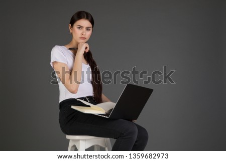 Upset unhappy businesswoman in white t-shirt leaning head on hand, sits on chair, looking at camera, holding book and laptop, posing in the studio on grey background