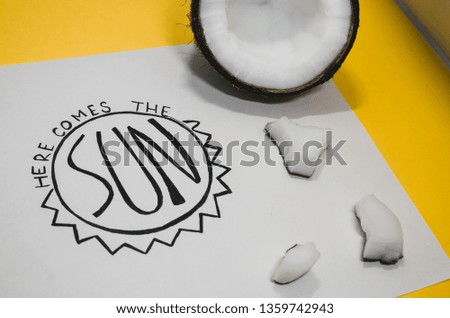 Hello summer concept.  A white paper where is wtitten with nice handwriting - Here comes the sun. Summer vibes and a half of ripe coconut on the yellow background