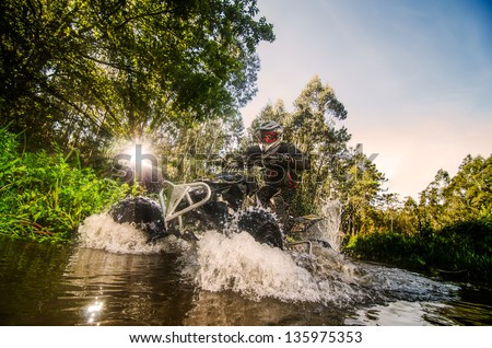Quad rider through water stream in the forest against sunlight.