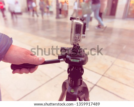 Male hand holding the handle of a tripod with an action camera on. Tripod is on a busy pedestrian street , a few people passing by in the background and a few stores blurry on purpose.