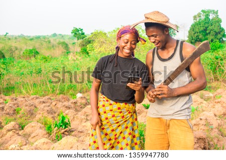 male and female African farmers on a farm smiling while viewing something on a phone together Royalty-Free Stock Photo #1359947780
