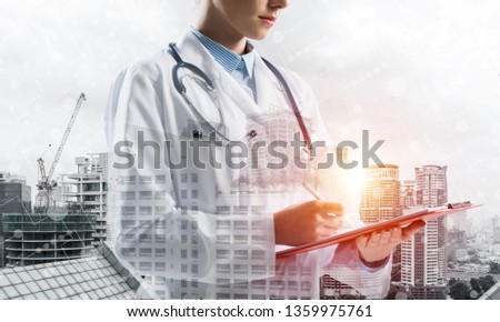 Side view of confident female doctor in white sterile coat making notes in notebook while standing outdoors with city view and medical interface icons on background. Medical industry. Double exposure