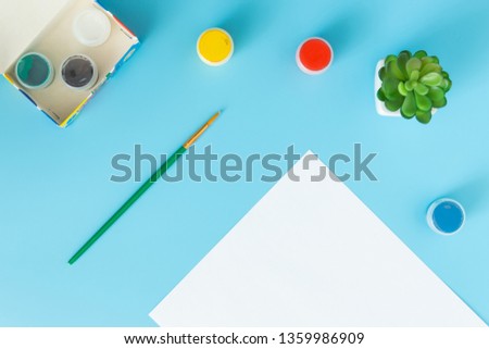 White sheet of paper with green succulent, multicolor paints and brush on a blue background with place for the inscription