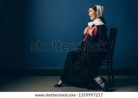 Try on new emotions or feelings. Medieval young woman as a nun in vintage clothing and white mutch sitting on the chair with bright pink bra on blue background. Concept of comparison of eras.