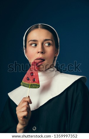 Keep it in secrets. Medieval young woman as a nun in vintage clothing and white mutch sitting on the chair and eating sweets on dark blue background. Concept of comparison of eras.