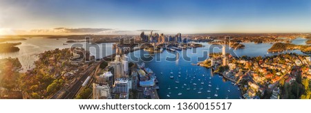Lavender bay from lower North Shore on Sydney harbour agains major city CBD landmarks around the Sydney Harbour bridge in soft warm morning light seen from mid-air over roof tops. Royalty-Free Stock Photo #1360015517