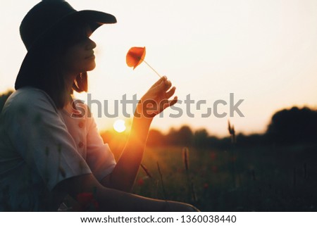 Silhouette of stylish girl in linen dress holding poppy flower in meadow in sun light with flowers in rustic straw basket. Boho woman in hat relaxing in summer sunset field. Atmospheric