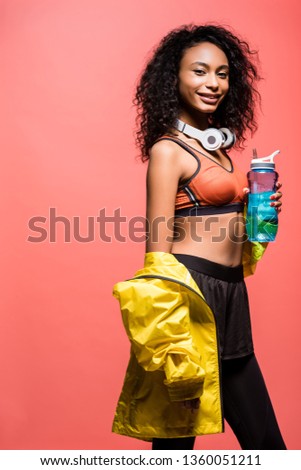 beautiful smiling african american sportswoman posing with headphones and sport bottle isolated on coral