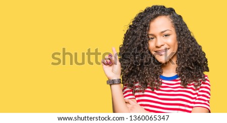 Young beautiful woman with curly hair wearing stripes t-shirt with a big smile on face, pointing with hand and finger to the side looking at the camera.