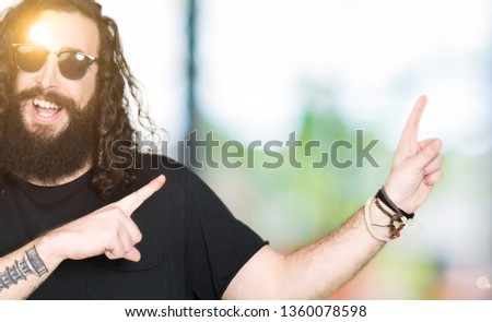 Young hipster man with long hair and beard wearing sunglasses smiling and looking at the camera pointing with two hands and fingers to the side.
