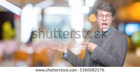 Young handsome business man wearing glasses over isolated background amazed and smiling to the camera while presenting with hand and pointing with finger.
