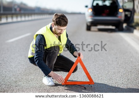 Man in road vest putting emergency triangle sign on the highway with broken car on the background