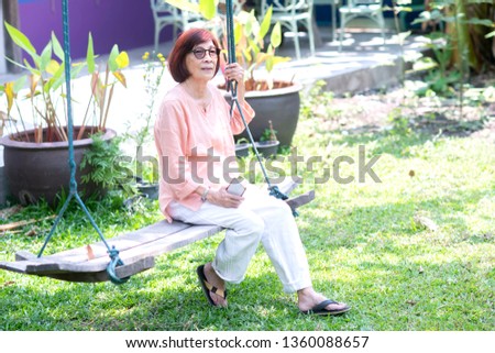 Asian senior woman sitting on the swing at  the outdoor garden  using smart phone to taking picture or face to face call, face recognition,on line chat. technology communication concept