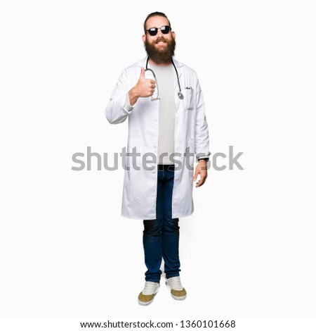 Young blond doctor man with beard wearing sunglasses doing happy thumbs up gesture with hand. Approving expression looking at the camera with showing success.