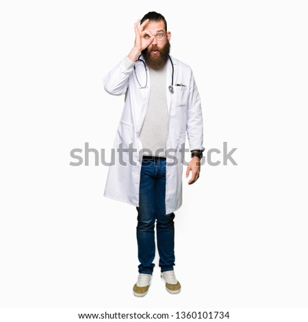 Young blond doctor man with beard wearing medical coat doing ok gesture shocked with surprised face, eye looking through fingers. Unbelieving expression.