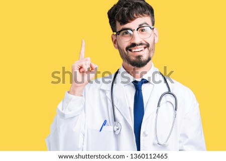 Young doctor man wearing hospital coat over isolated background pointing finger up with successful idea. Exited and happy. Number one.