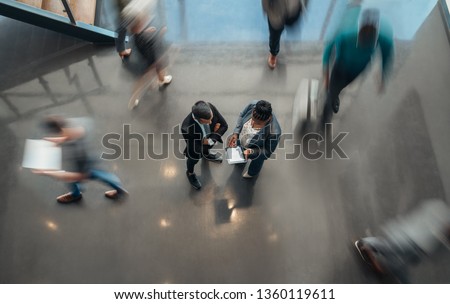 Two business people standing in the lobby of an office looking at a tablet while people are walking past in a blur