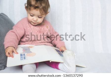 Little baby girl reading a picture book on a white sofa