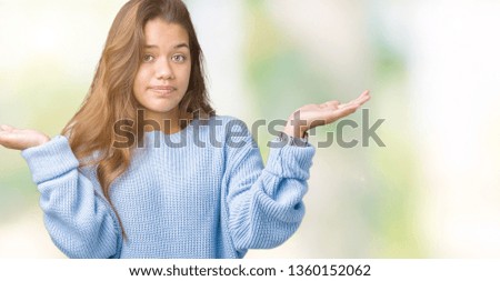 Young beautiful brunette woman wearing blue winter sweater over isolated background clueless and confused expression with arms and hands raised. Doubt concept.