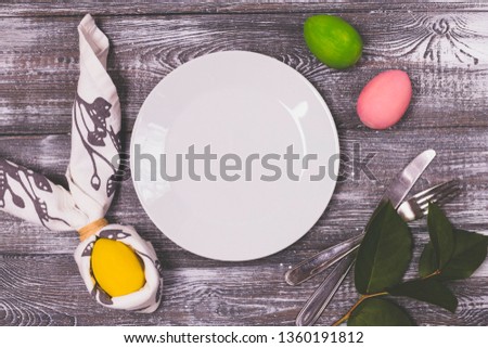 Top view of a Easter egg wrapped in a napkin in the shape of rabbit ears, a white plate and cutlery, colorful Easter eggs on a wooden gray table.
