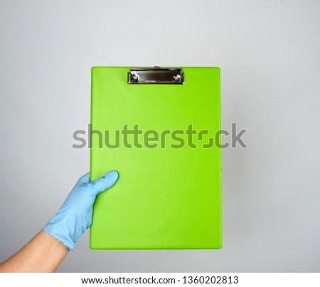 female hand with blue medical glove holding a green tablet for clamping papers on gray background
